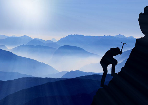 Das Bild zeigt aus ca. 10 Meter Entfernung einen Bergsteiger vor einer blau eingefärbten Bergkulisse. Er scheint gerade im rechten Bildrand seinen Gesteinspickel in den Berg zu schlagen.