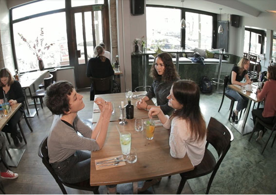 Das Bild zeigt eine Szene auf einem der AVWS-Videos. Es sitzen drei junge Damen an einem Tisch in einem Restaurant. Verteilt an vereinzelten umliegenden Tischen sitzen weitere Personen im Restaurant.